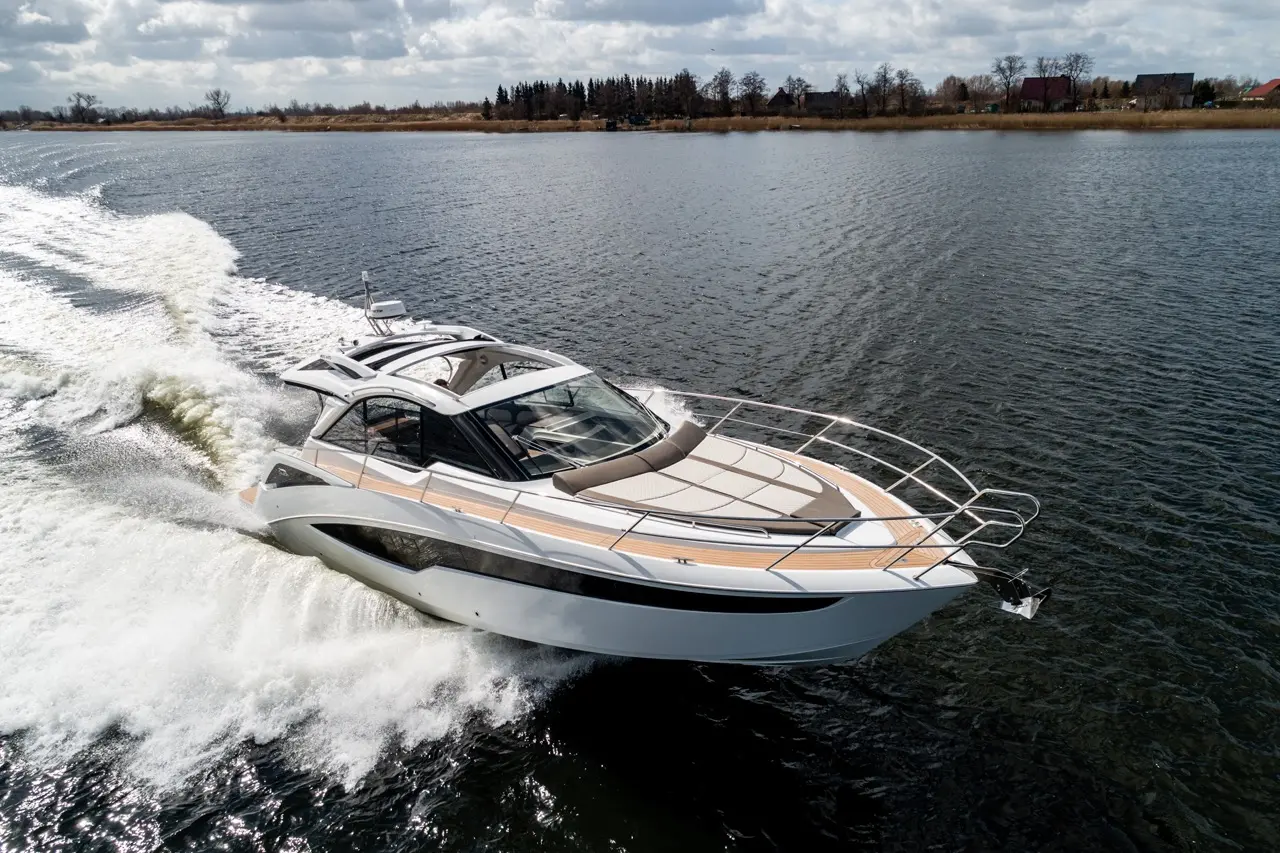 A sleek motorboat speeds across calm waters, leaving a white wake, with trees and buildings on the distant shore under a cloudy sky.