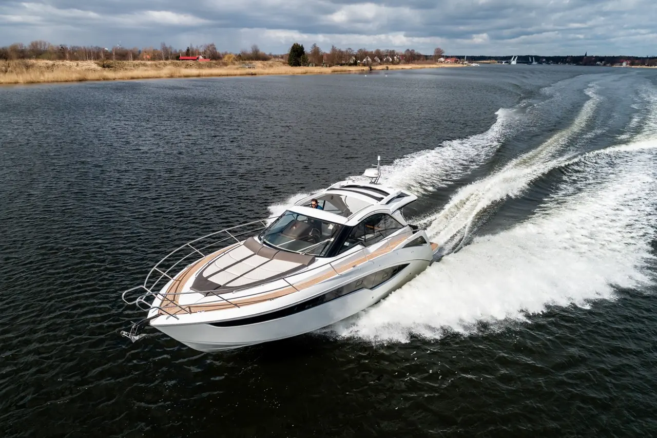 A sleek white motorboat navigates through a dark river, creating spray under a cloudy sky. Scenic shoreline visible.