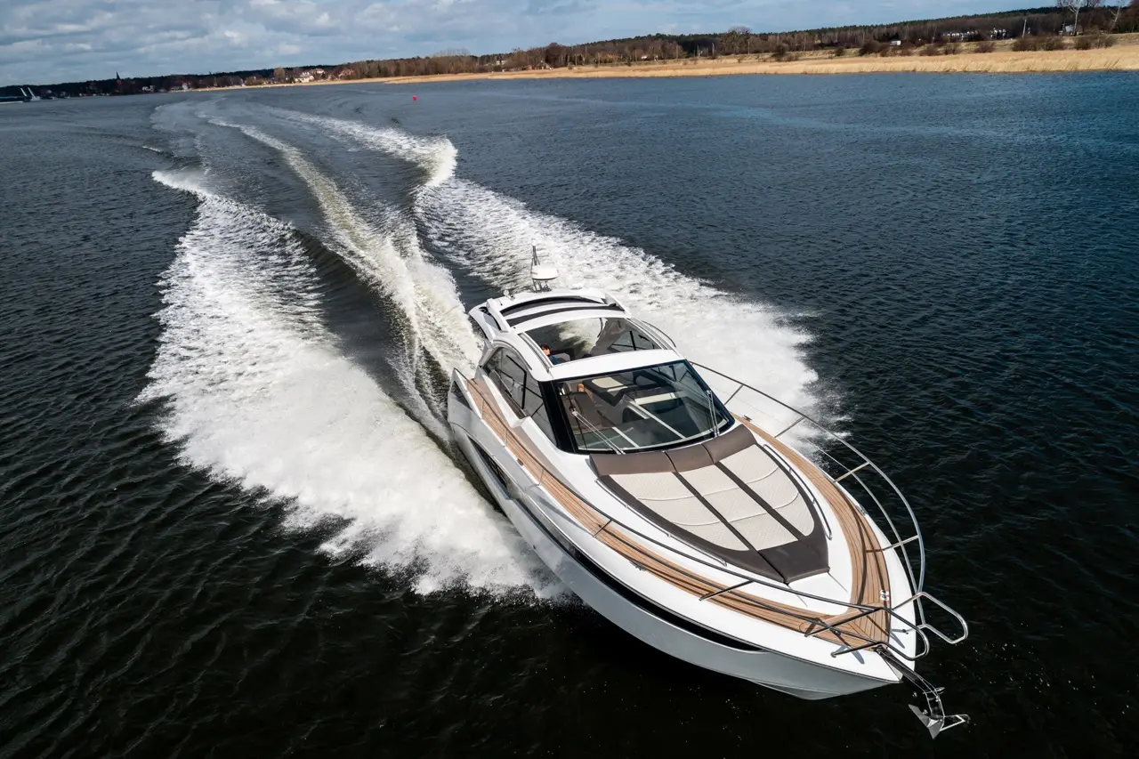 A sleek motorboat speeds across calm waters, leaving a white wake against a serene landscape under a partly cloudy sky.