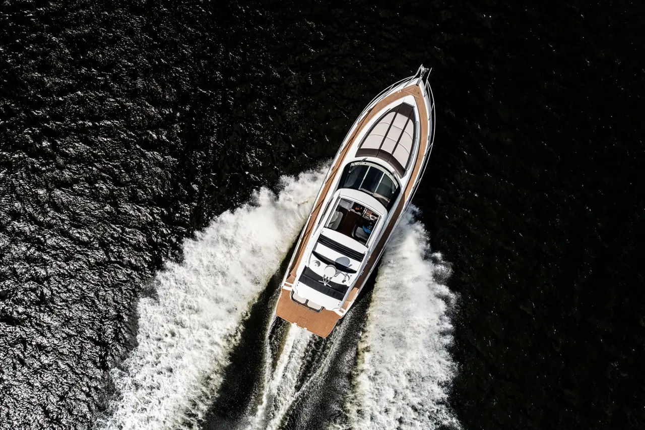 A luxury motorboat speeds across dark water, creating a white wake in its path under bright sunlight.