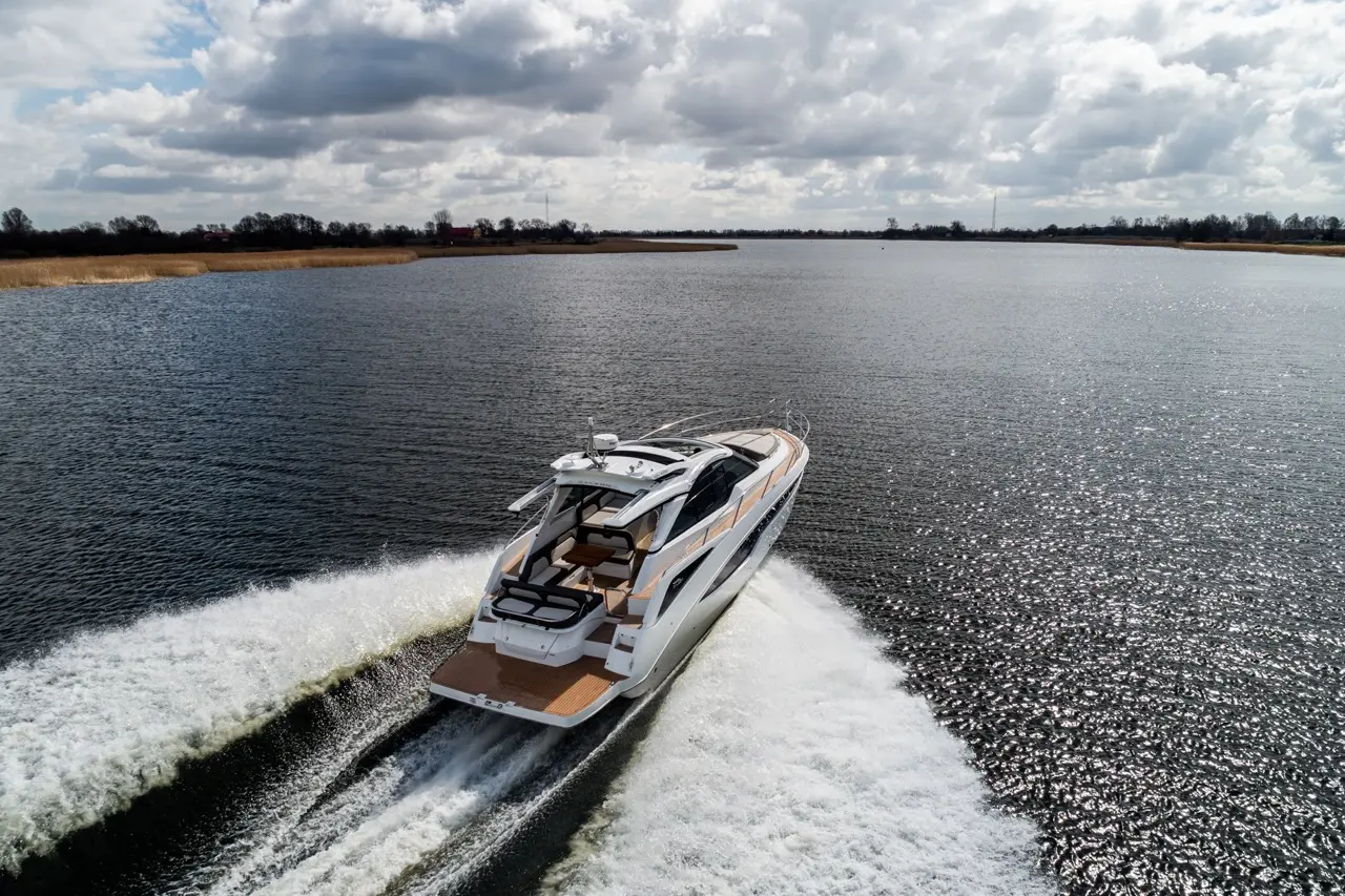 Luxury motor yacht gliding across a sparkling river under a cloudy sky, leaving a frothy wake behind.