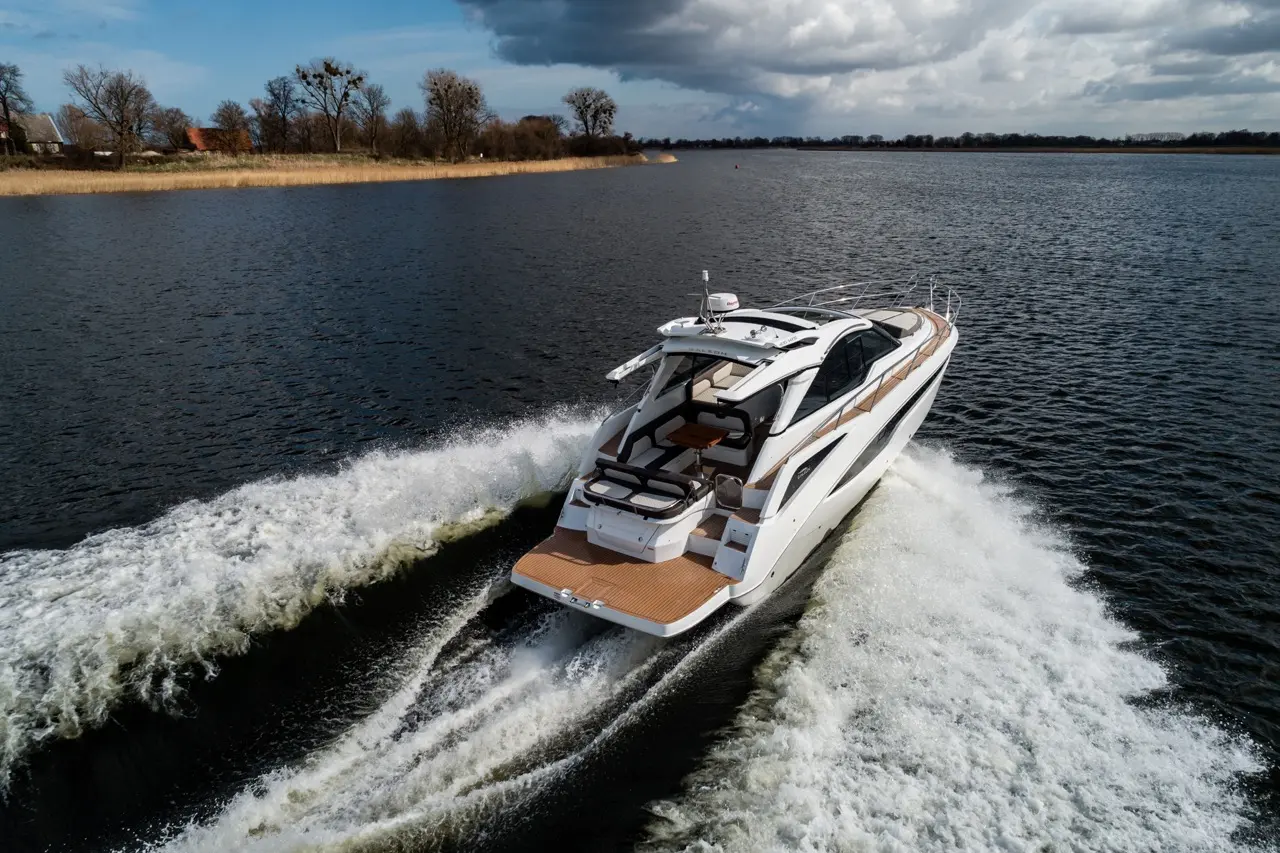 A luxury motorboat cruising on a lake, creating white foamy waves against a backdrop of trees and cloudy skies.
