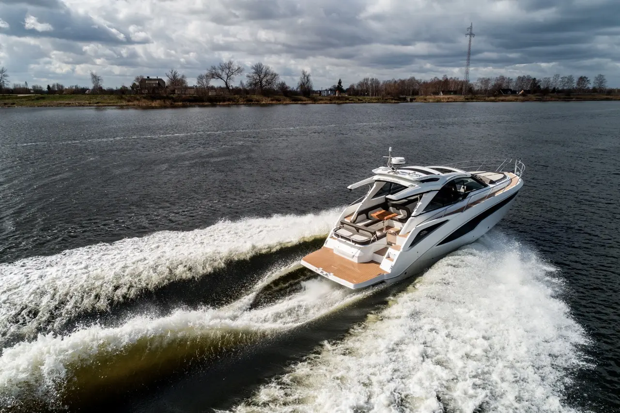 A sleek yacht gliding across a river, leaving a wake, under a cloudy sky with trees and a distant house on the shore.