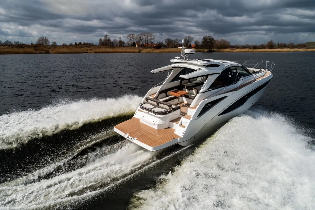 A luxury motor yacht accelerates on a calm lake, leaving a wake and surrounded by overcast skies and distant shorelines.