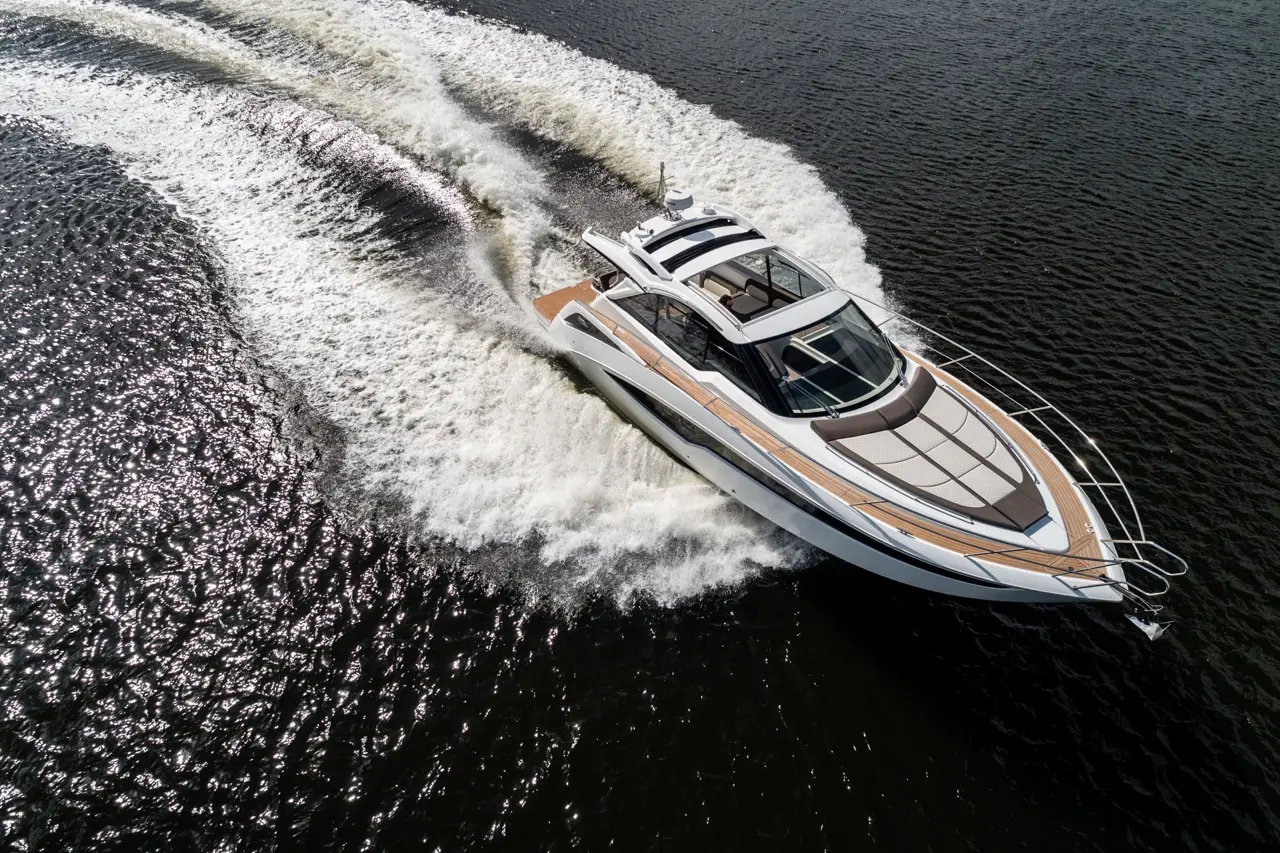 A sleek white yacht with wooden detailing speeds through dark water, creating a wake under bright sunlight.