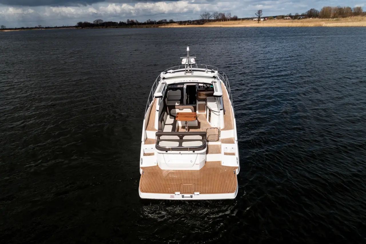 Aerial view of a sleek motorboat on calm water, featuring a spacious deck with seating and a wooden table, under a cloudy sky.