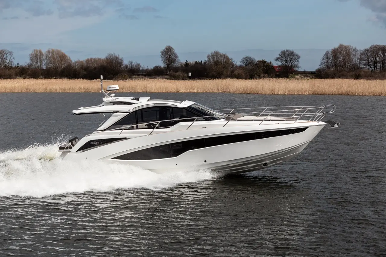 A sleek white motorboat speeds through a calm waterway, with reeds and trees visible in the background.