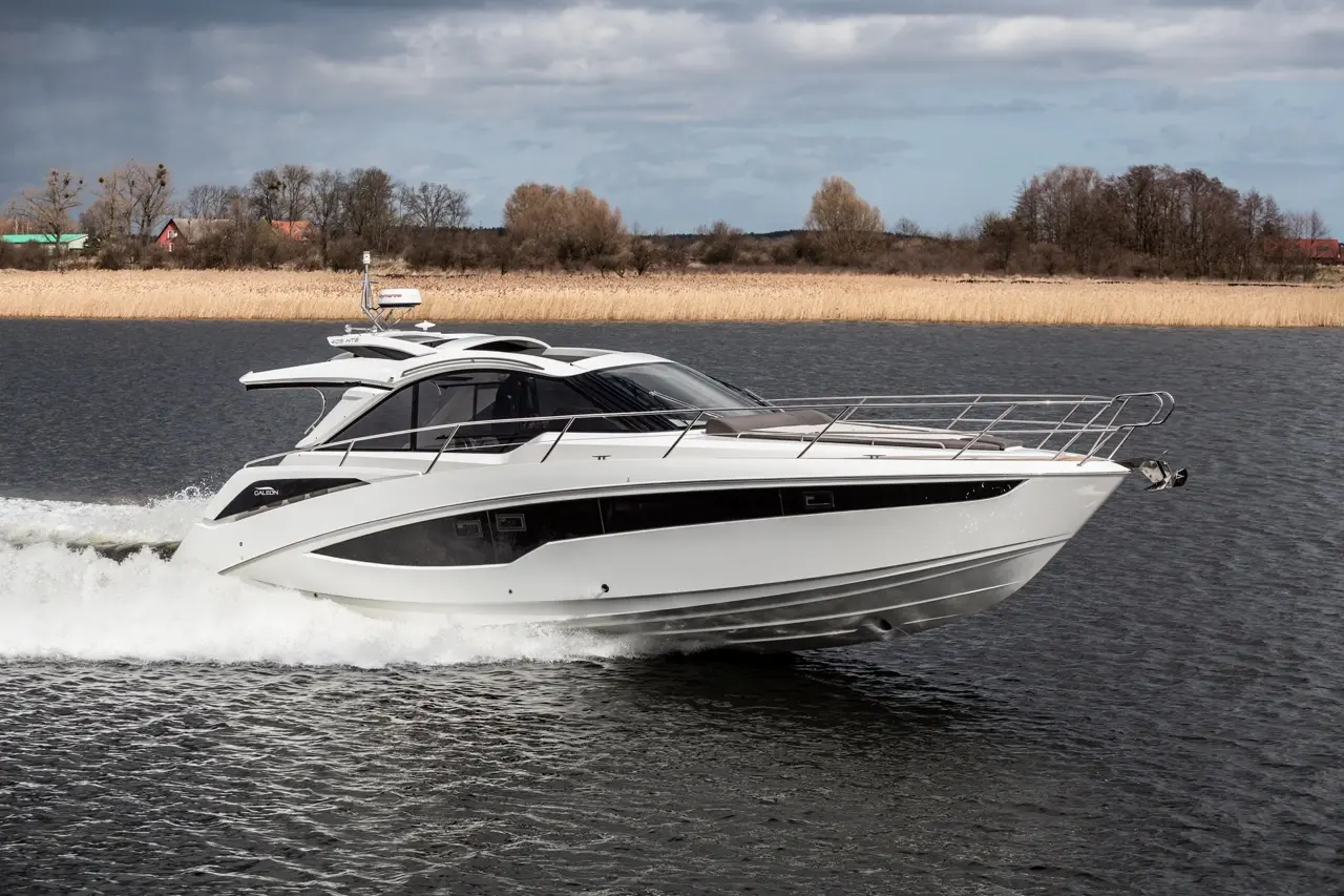 A sleek white motorboat cruises through calm waters, creating a spray, with trees and a golden reeds backdrop.