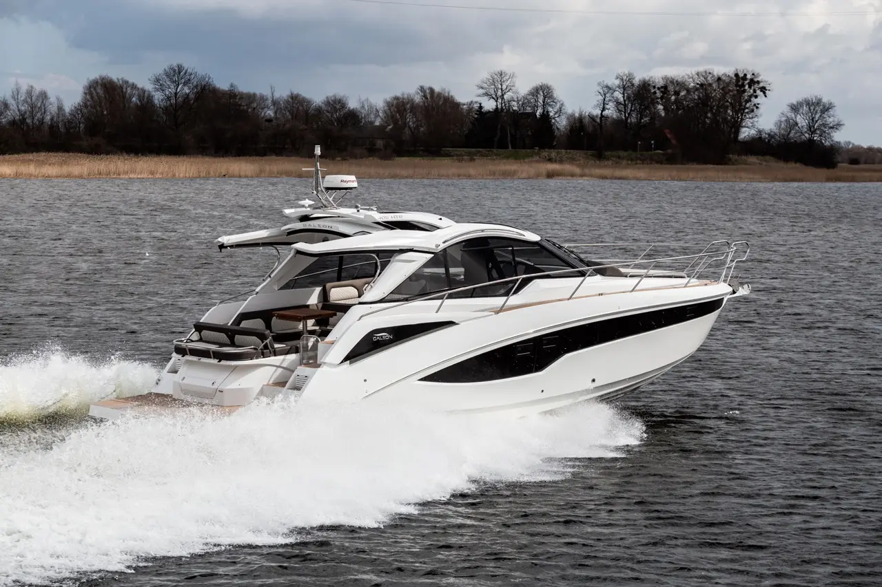 A sleek white motorboat speeds across a lake, creating a spray of water against a backdrop of trees and cloudy skies.