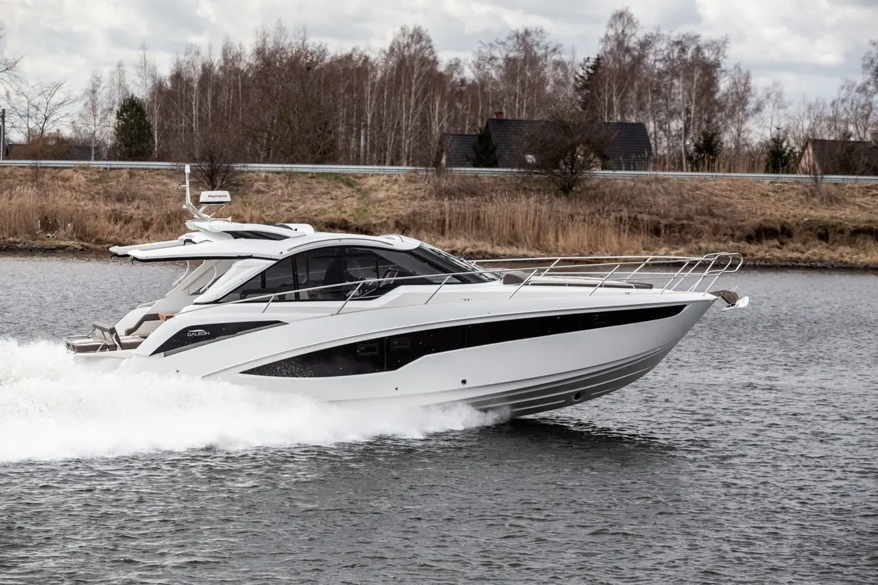 A sleek white motorboat speeds across a calm river, with trees and a house in the background, showcasing a modern design.