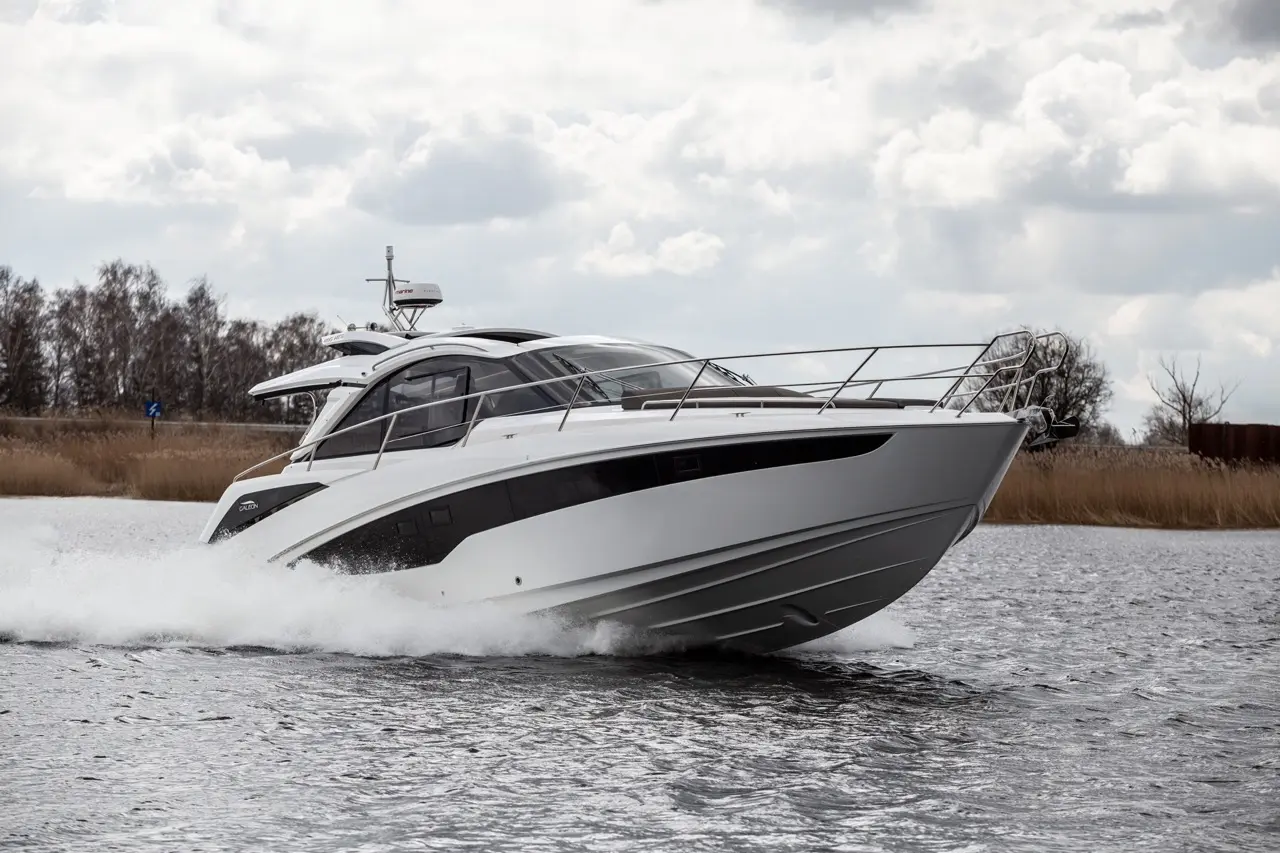 A sleek white motor yacht accelerates through calm waters, creating splashes, under a cloudy sky.