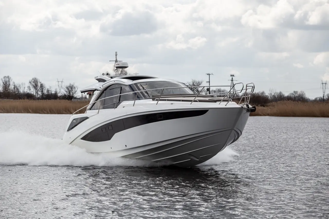 A sleek white motorboat powering through water, creating waves, with a cloudy sky and distant trees in the background.