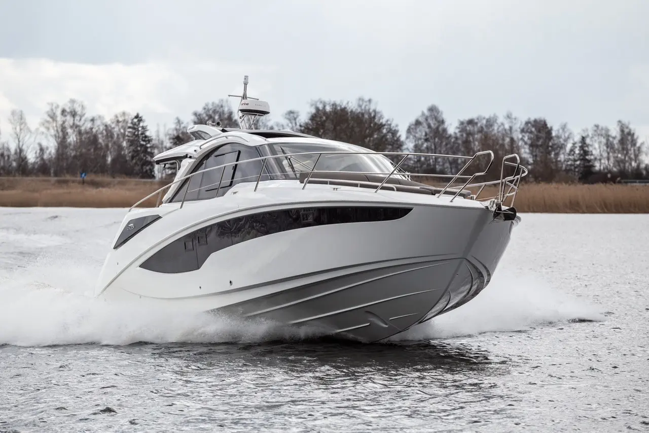 A sleek motorboat accelerates across water, creating splashes, with trees and a cloudy sky in the background.