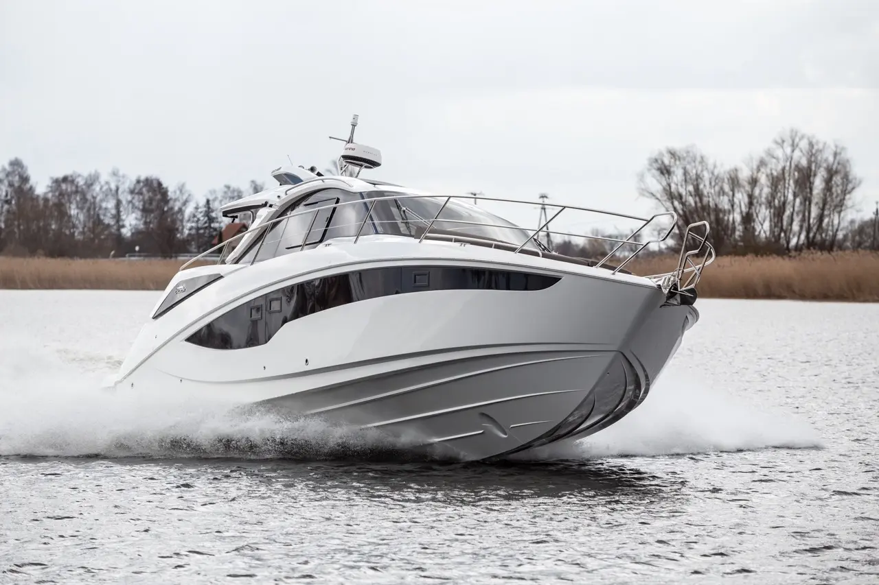 A sleek white motorboat skimming across the water, creating splashes, with trees and reeds visible in the background.