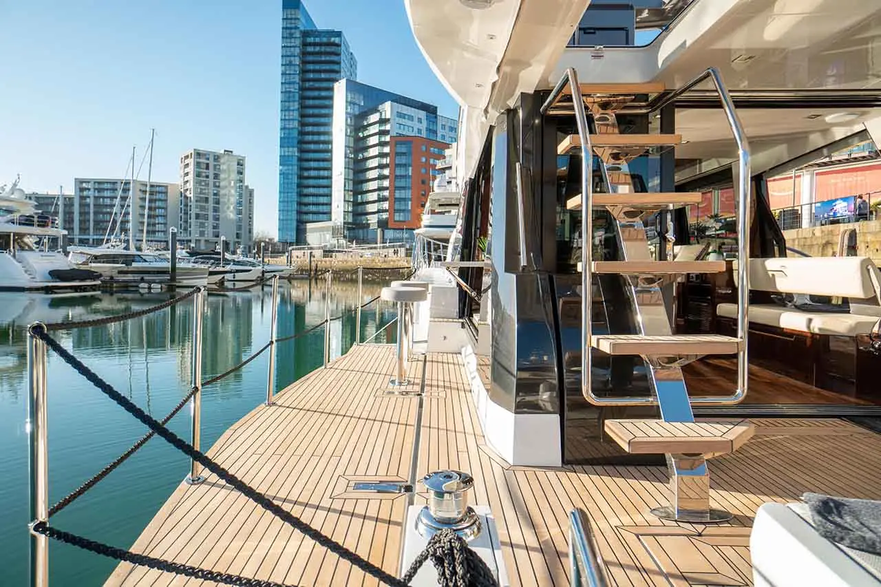 Luxury yacht docked, featuring a stylish wooden deck, modern stairway, and views of contemporary buildings and boats.
