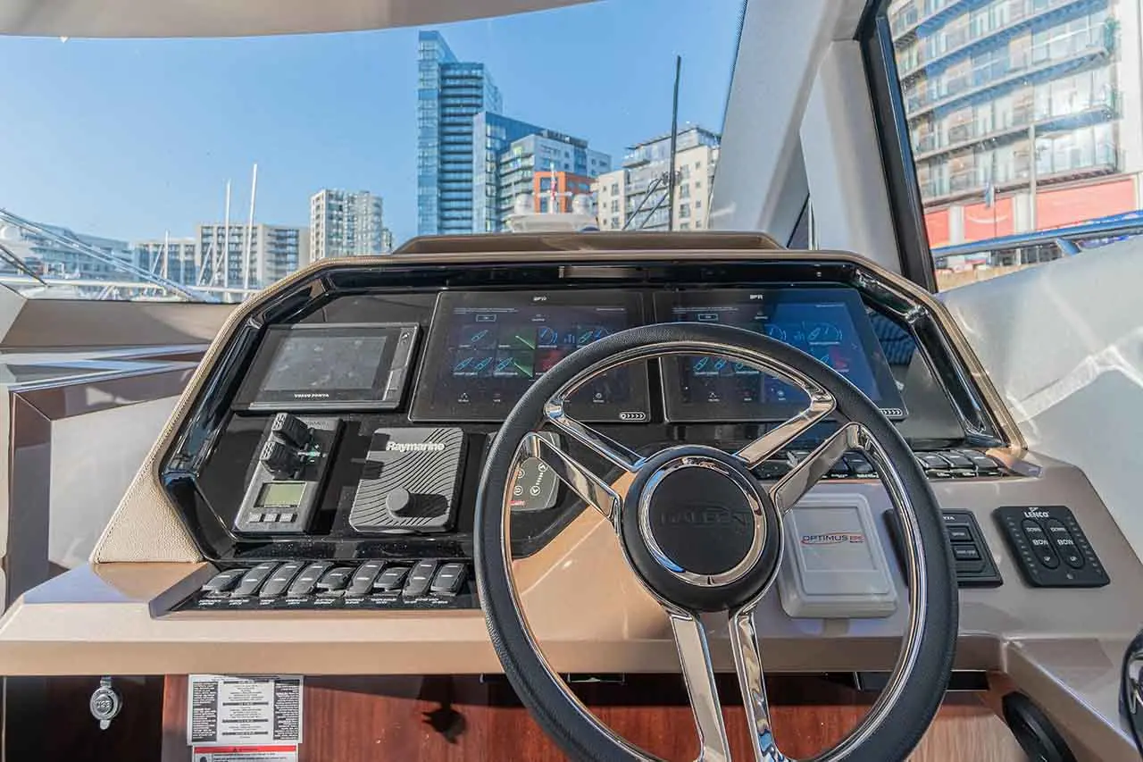 Boat dashboard with multiple screens displaying navigation data, steering wheel, and urban buildings visible through the window.