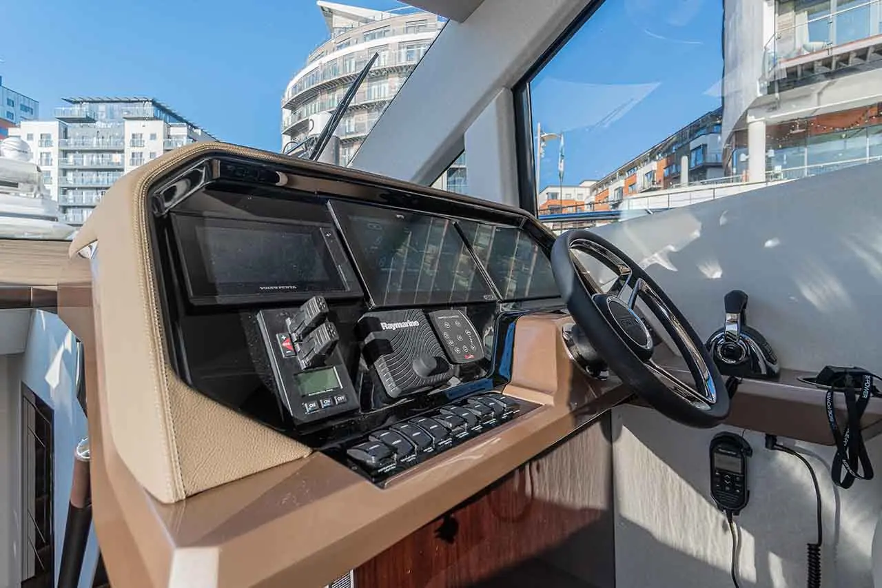 Modern boat helm featuring dual screens, navigation controls, and a steering wheel overlooking a marina with buildings.