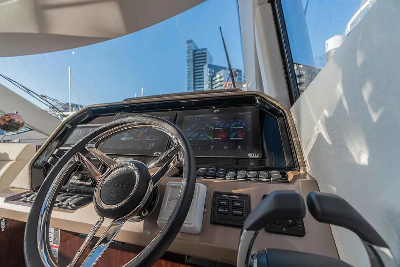Boat helm featuring a steering wheel, digital displays, and control buttons, with a backdrop of city buildings and clear blue sky.