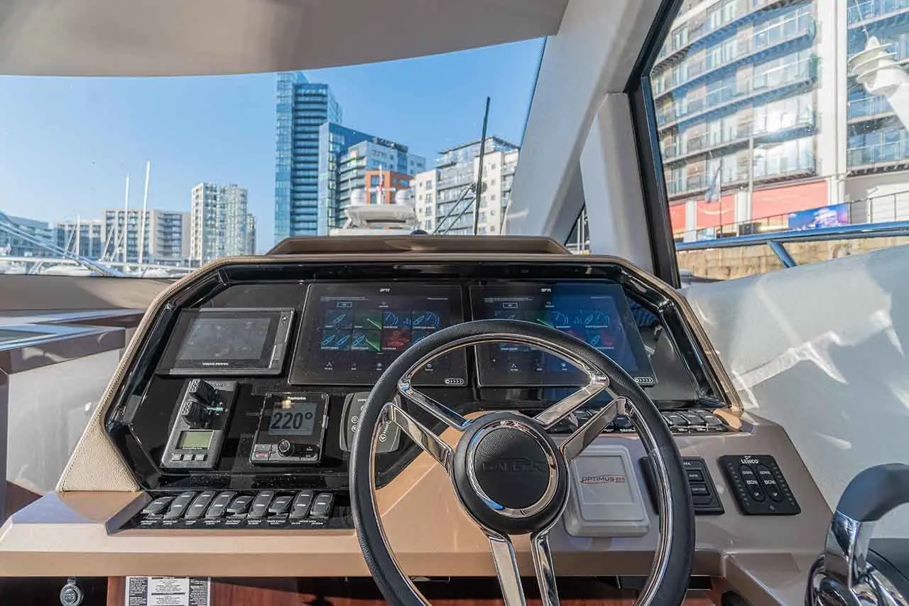 Boat's control cockpit with steering wheel and digital displays, showcasing navigation data and urban skyline in background.