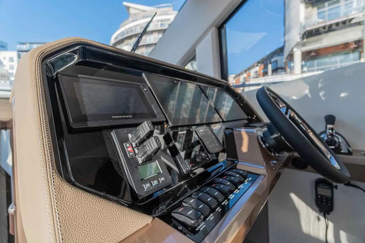 Boat dashboard featuring control panels, displays, and switches, with a view of modern buildings through large windows.
