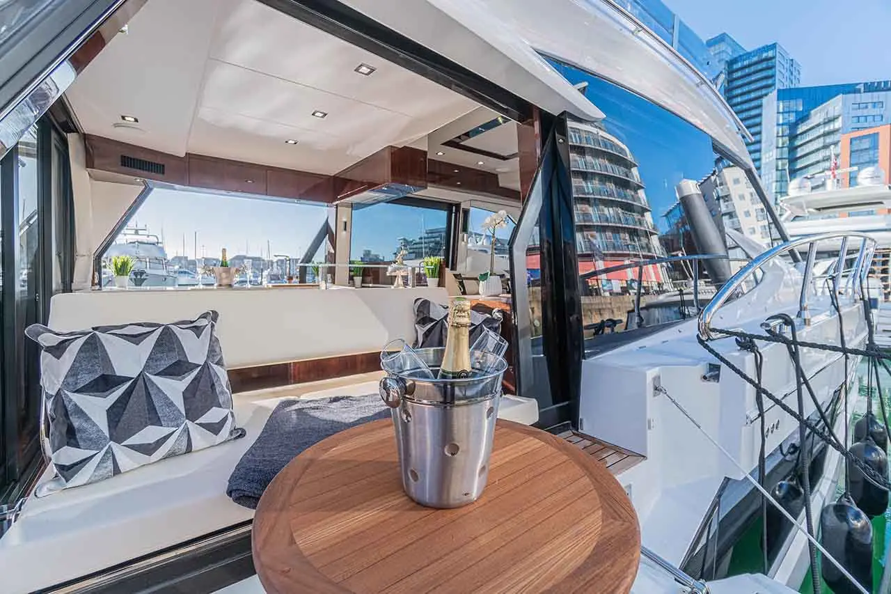 Luxury yacht interior featuring a polished wood table, champagne bucket, geometric cushions, and marina views.