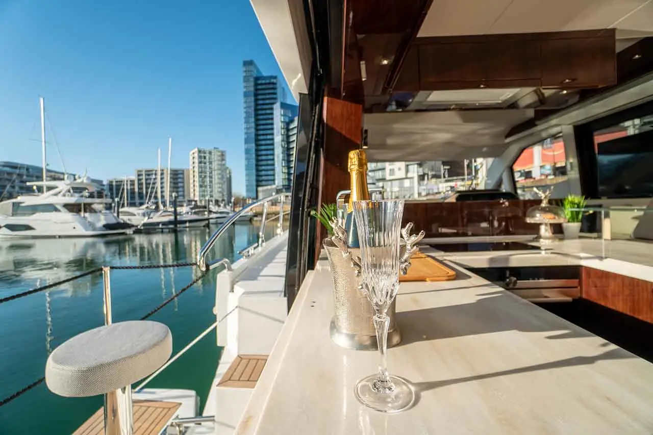 Luxury yacht interior with a champagne bottle, crystal glass, and marina view featuring modern buildings and boats.