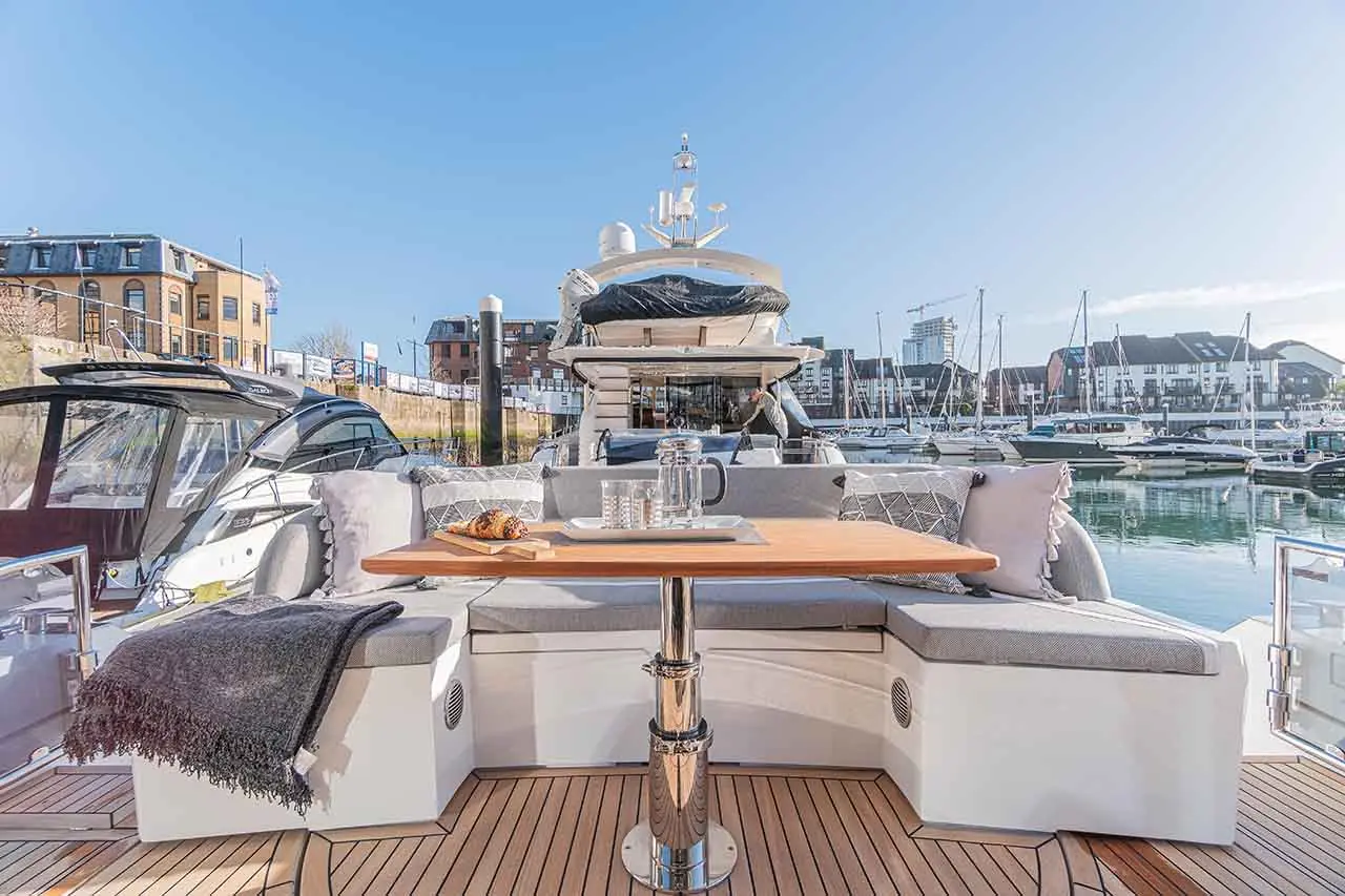 Elegant boat interior featuring a wooden table, grey cushioned seating, coastal views, and a glass jug with cups.