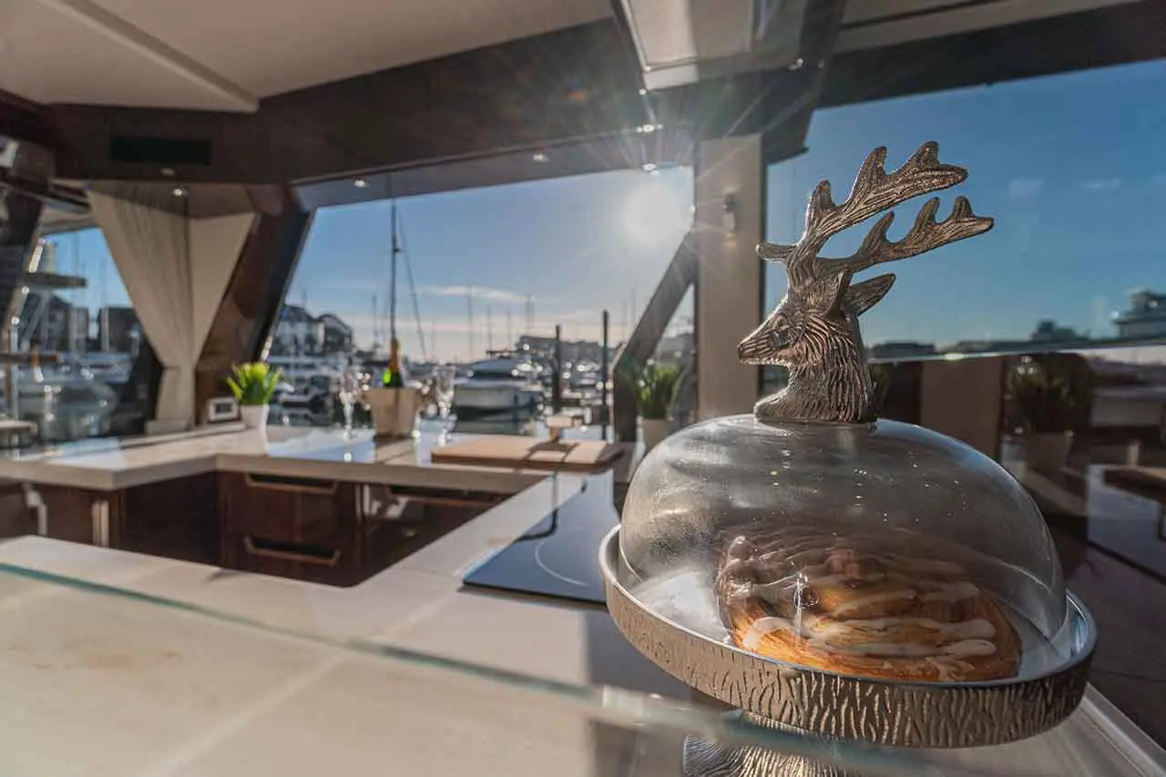 Stylish yacht interior with a pastry under a glass dome, a reindeer decor, and a marina view through large windows.