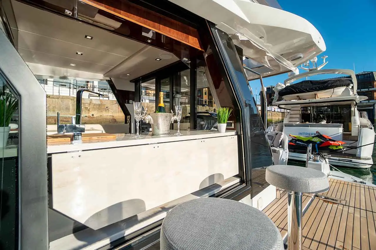 Luxury yacht interior featuring a bar with champagne, glasses, and modern décor, alongside water and nearby boats.