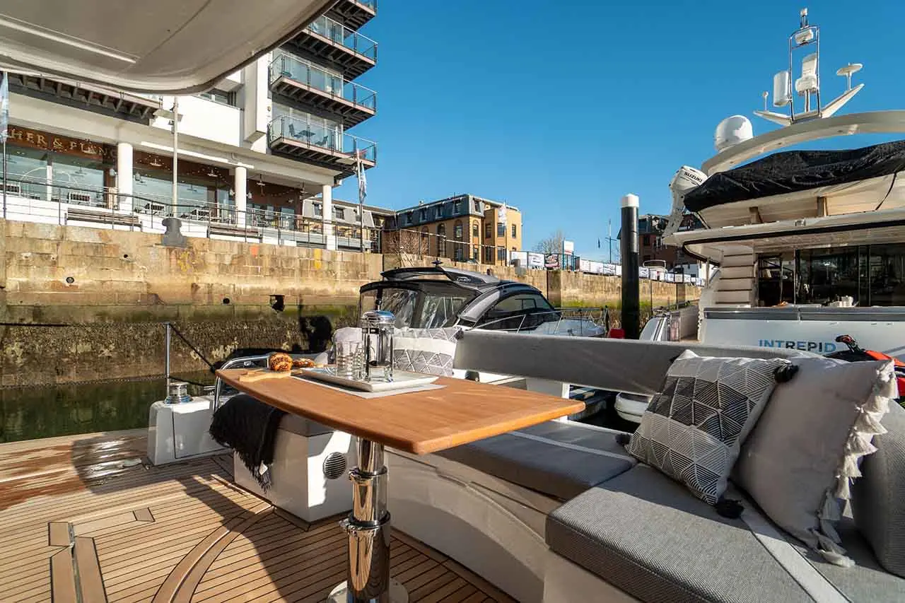 A stylish boat deck features a wooden table set with glassware, cushions, and a view of nearby buildings and boats.