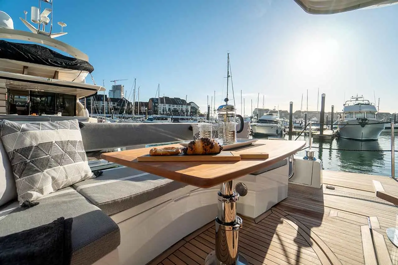 A sunny marina view featuring a wooden table with a croissant and glasses, surrounded by boats and modern buildings.