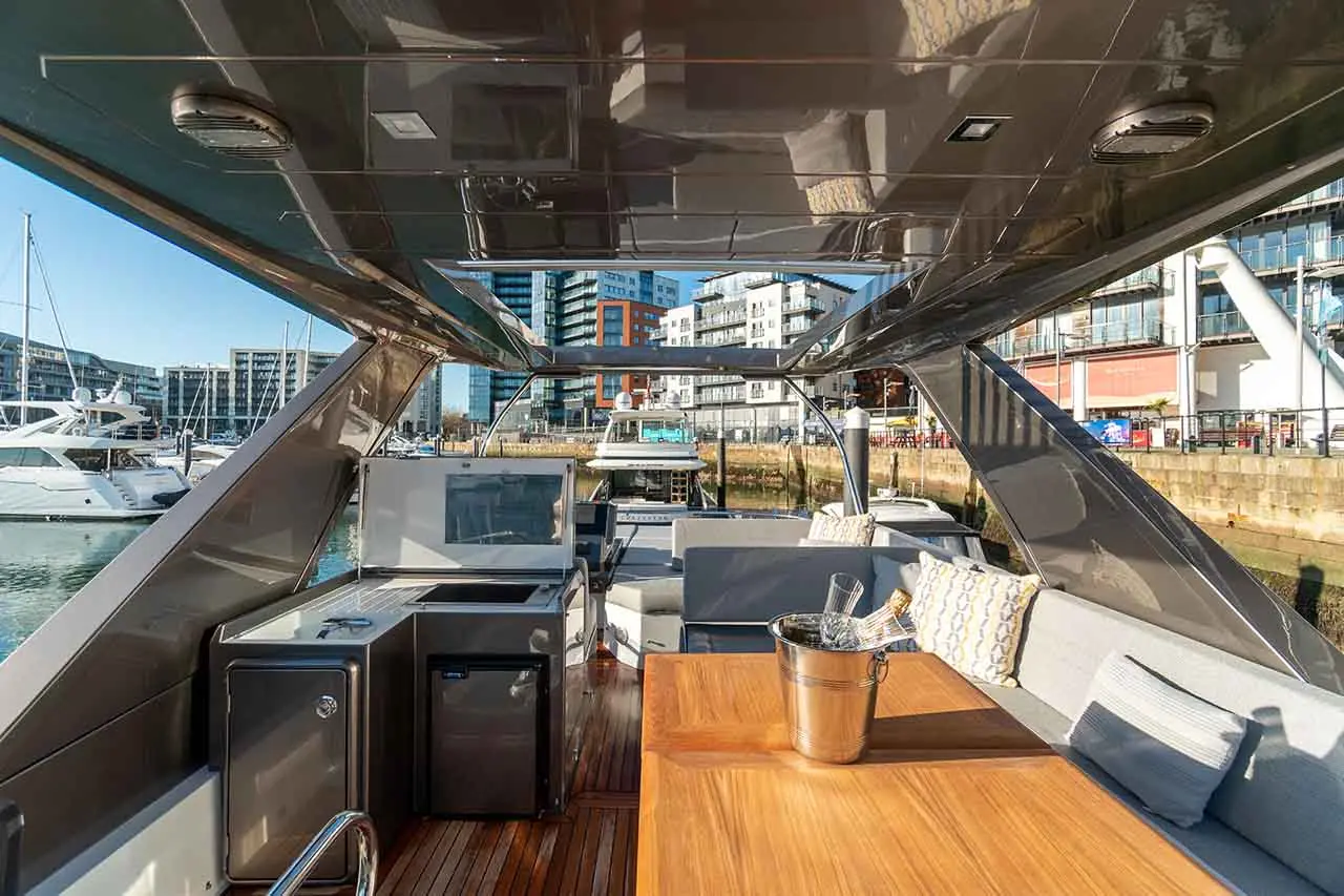 View of a luxury yacht interior with a wooden table, seating, and an ice bucket, overlooking modern marina buildings.