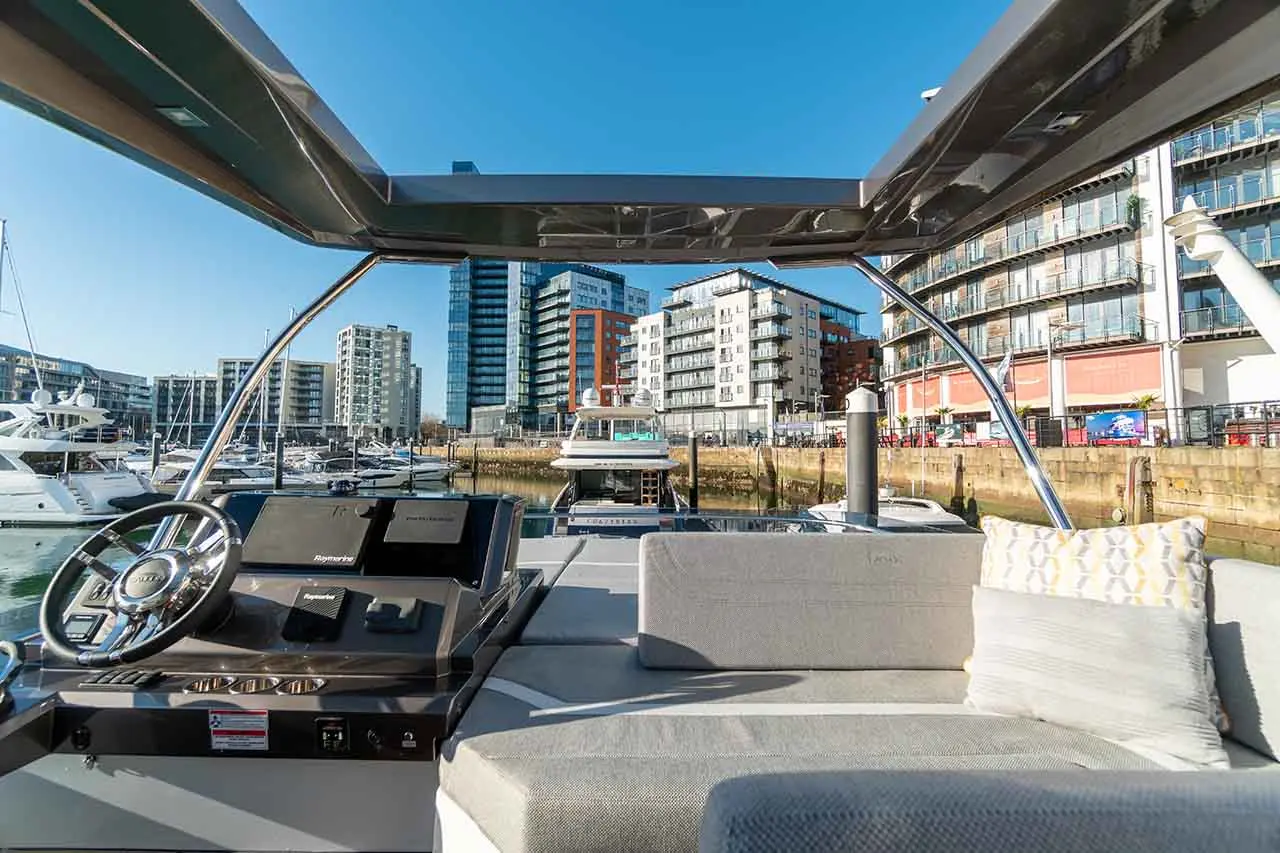 View from a boat's cockpit showing the steering wheel, instrument panel, and modern buildings in the marina backdrop.