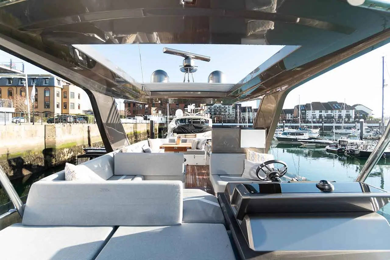 Luxury yacht interior with grey seating, wooden table, and helm, overlooking a marina filled with boats and modern buildings.