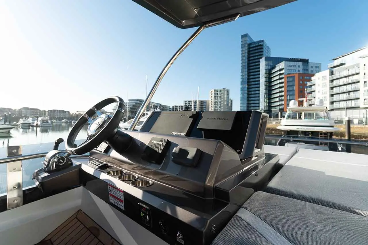 Boat cockpit view with steering wheel, control panel, and modern buildings in the background, reflecting a marina setting.
