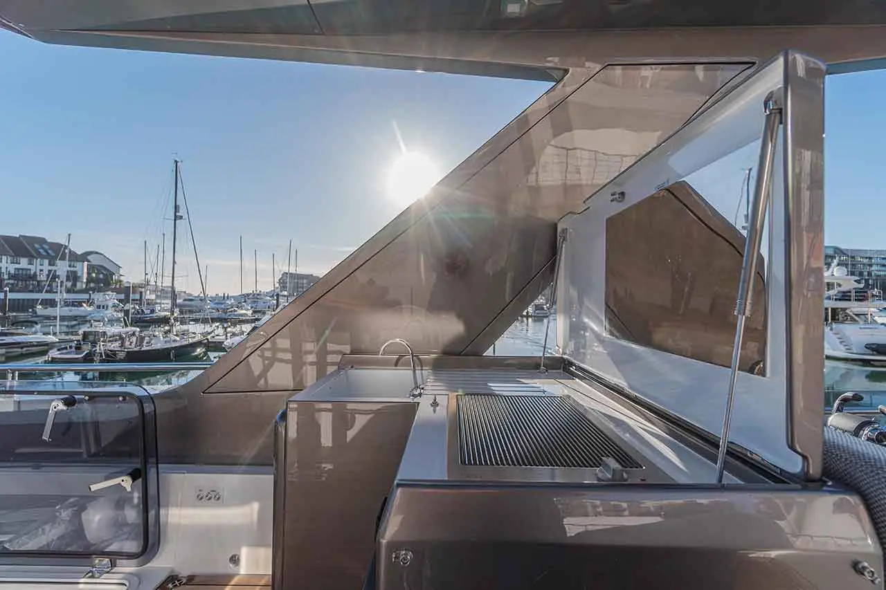 View from a yacht's deck featuring a stainless-steel grill and sink, with a marina and boats illuminated by the sun.