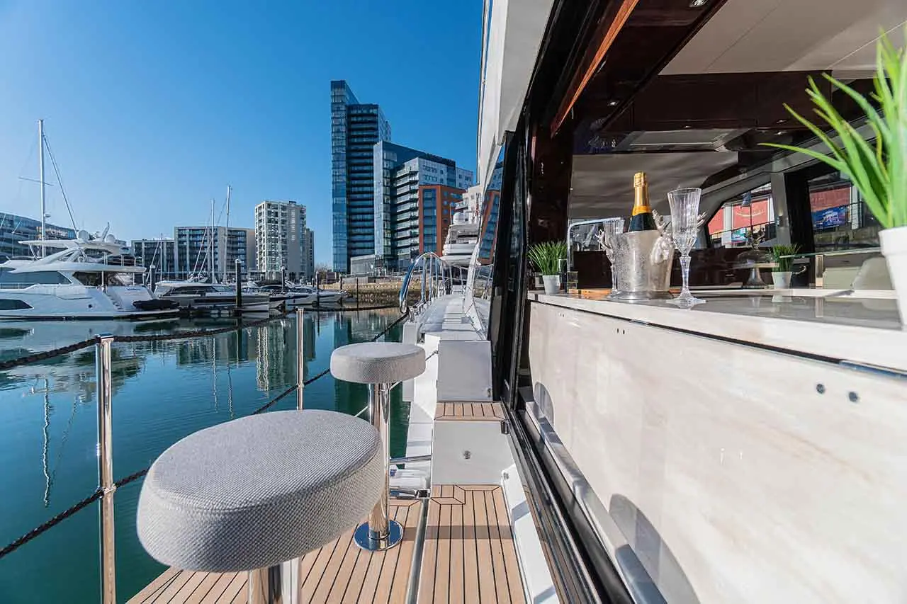 Modern yacht with stylish bar, champagne, and a view of a marina and city buildings under a clear blue sky.