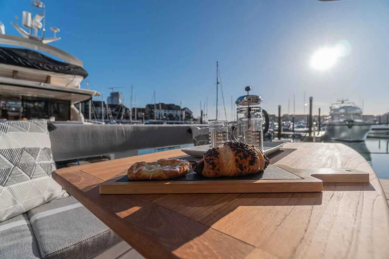 Breakfast on a wooden table with a chocolate croissant and pastry, overlooking a marina with boats and a bright sun.