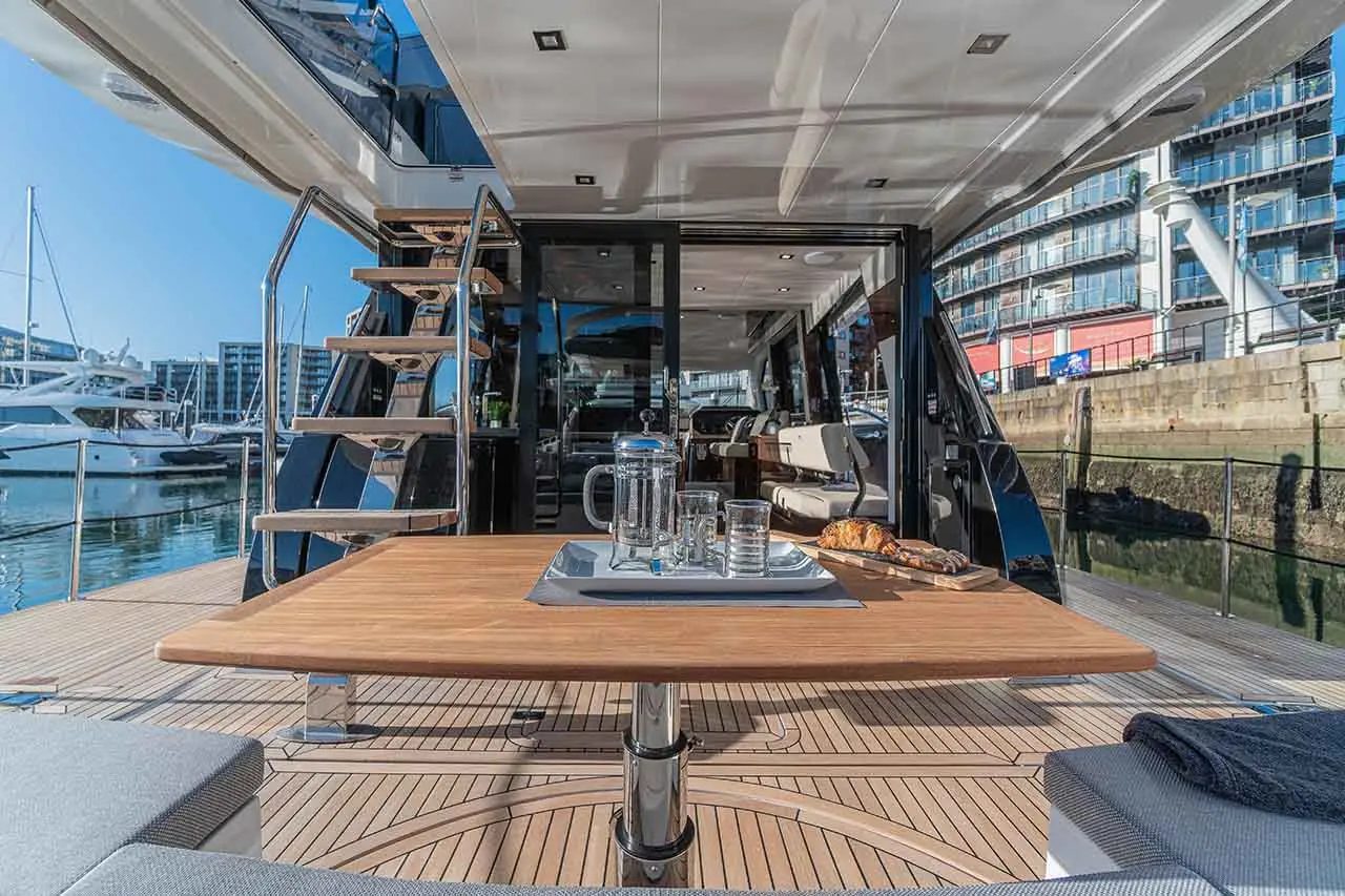 Luxurious yacht deck with a wooden table set for breakfast, featuring glassware and pastries, against a sunny marina backdrop.