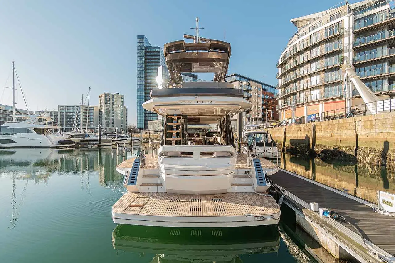 A luxury yacht docked at a marina, with modern buildings and reflections in the calm water under a clear blue sky.