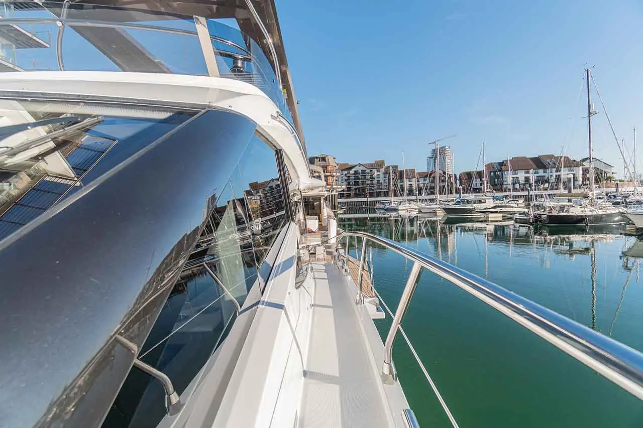 A yacht's deck overlooks a marina filled with boats, reflecting buildings and blue sky on calm waters.