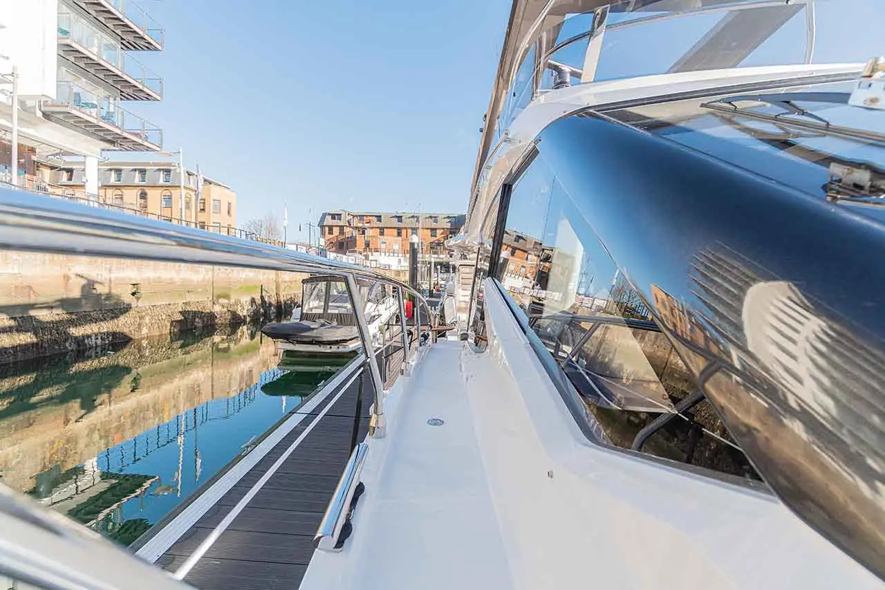 View from a yacht's deck showing gleaming railings and reflections, with moored boats and waterfront buildings in the background.