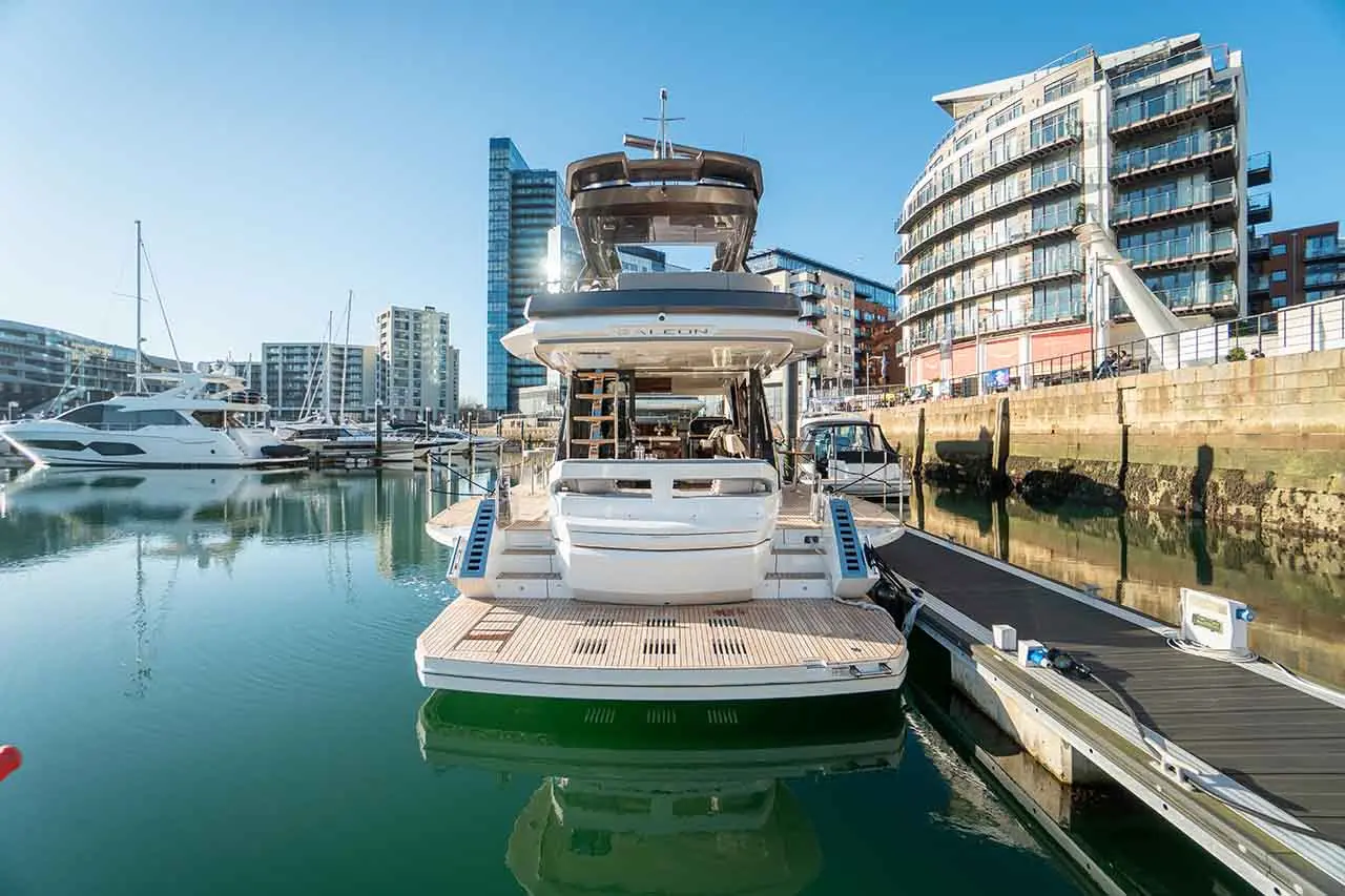Luxury yacht docked in a marina, surrounded by modern buildings and calm water reflecting the clear blue sky.