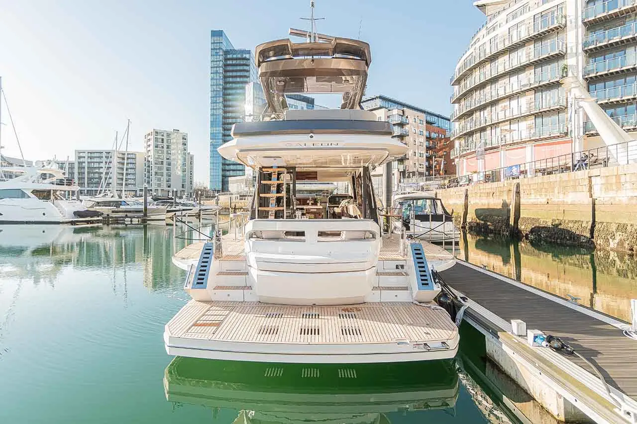 Luxury yacht docked in a marina with modern buildings in the background, reflecting calm waters under a clear sky.