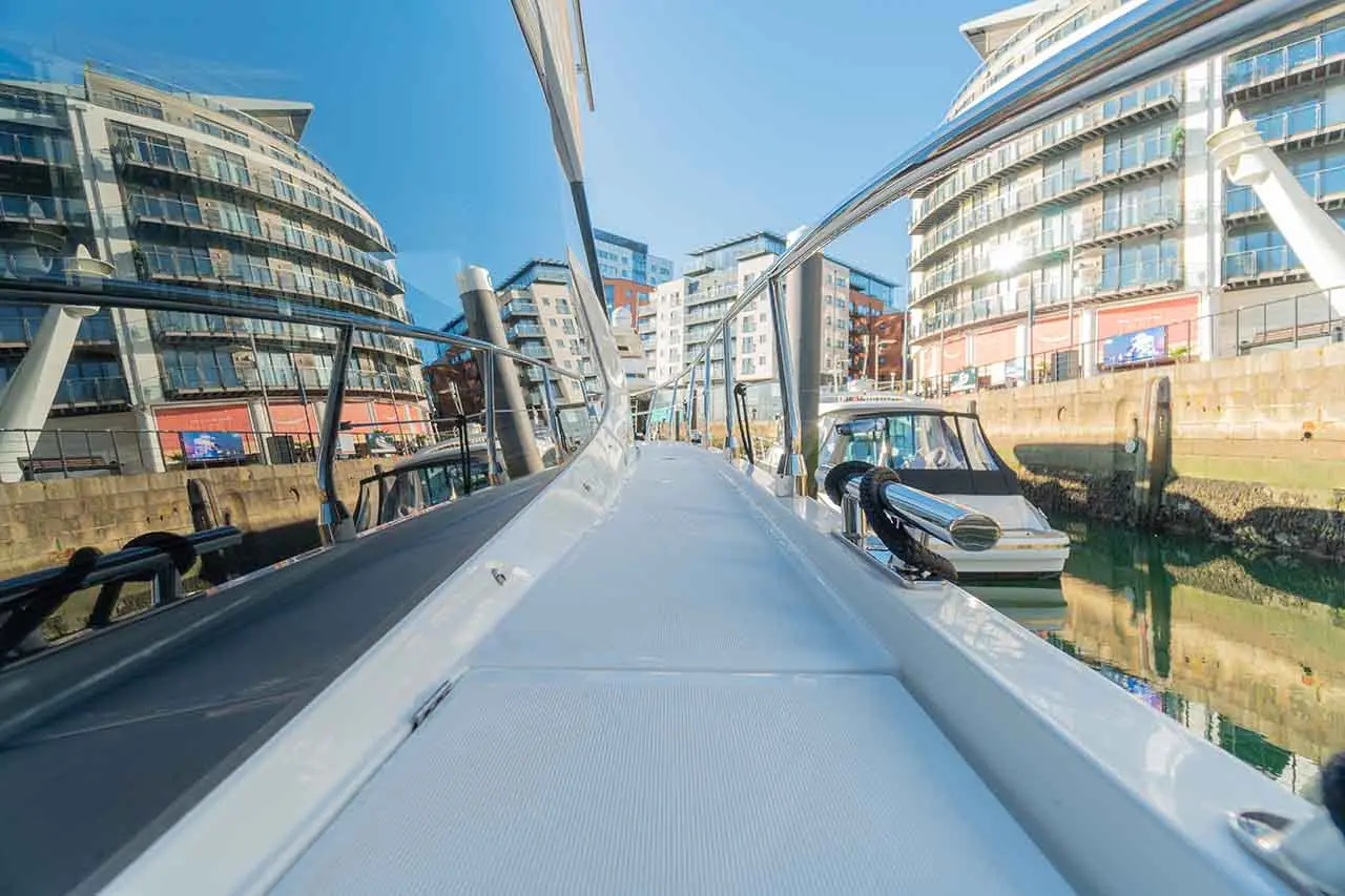 View from a boat deck, reflecting modern buildings and boats in a marina under a clear blue sky.