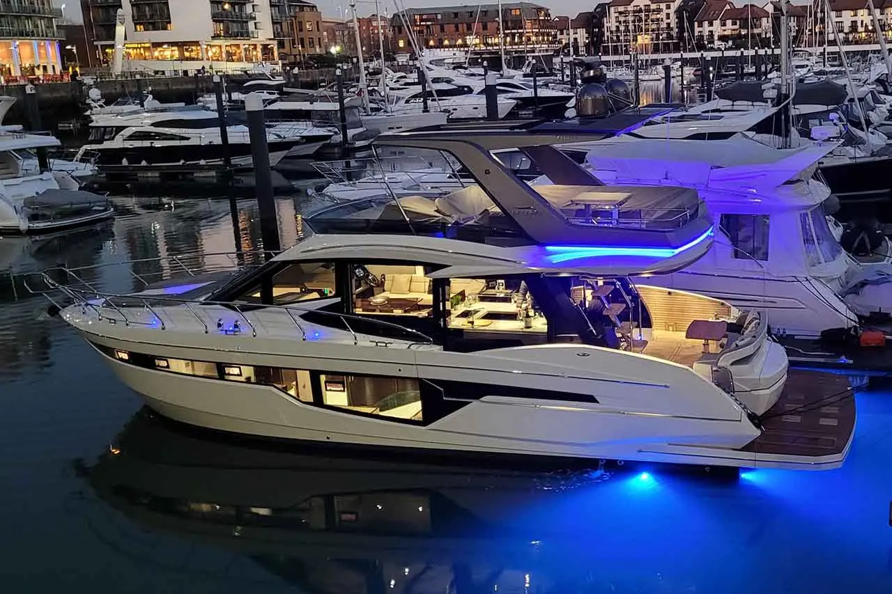 Luxurious motor yacht at a marina, illuminated with blue lights, surrounded by boats and buildings in the evening.