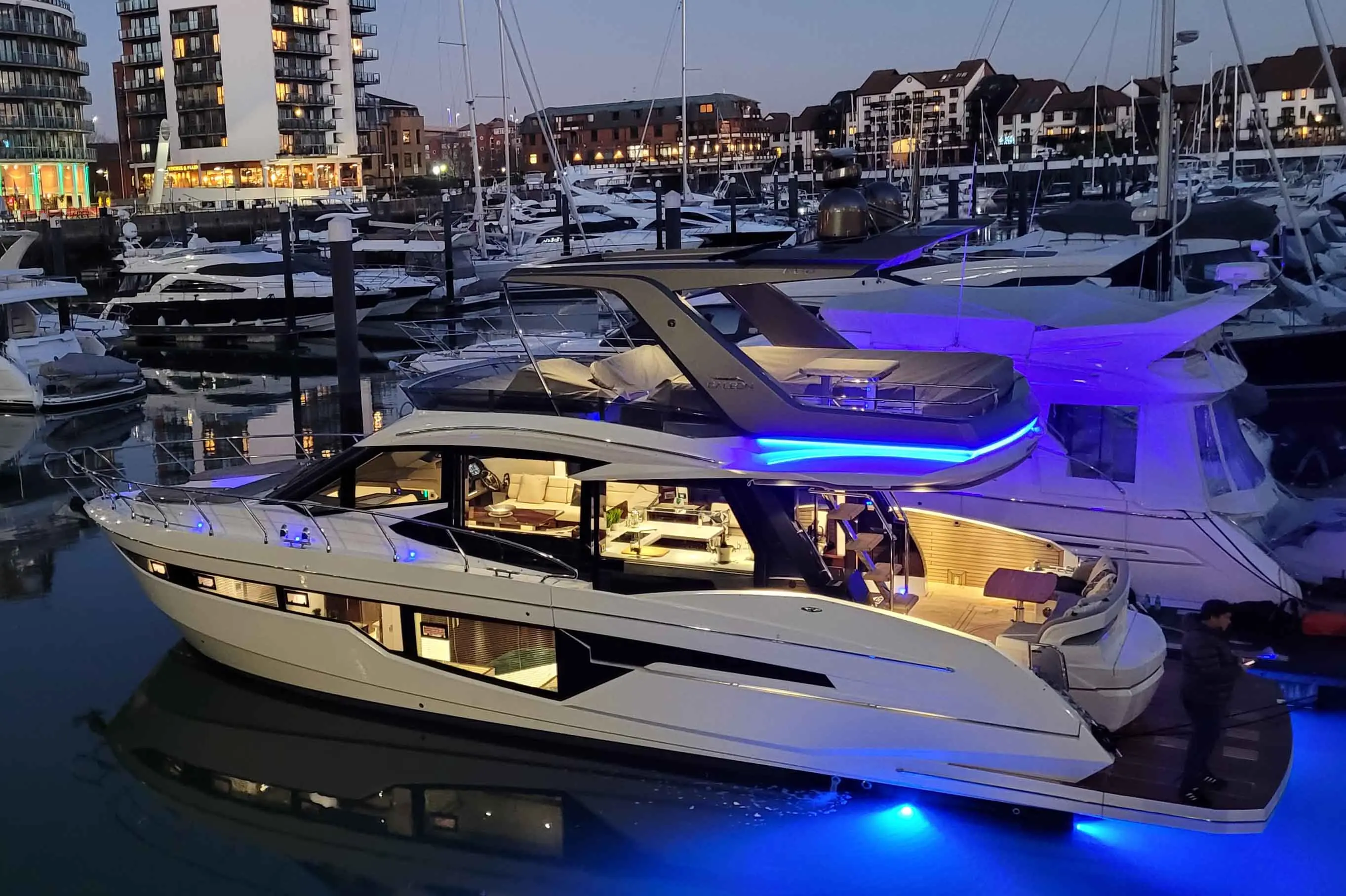 Luxury motor yacht with illuminated features docked in a marina, surrounded by other boats and waterfront buildings at dusk.