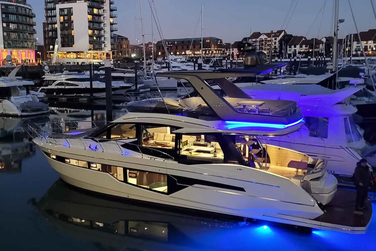 A luxury yacht illuminated by blue lights docked at a marina, surrounded by other boats with buildings in the background.