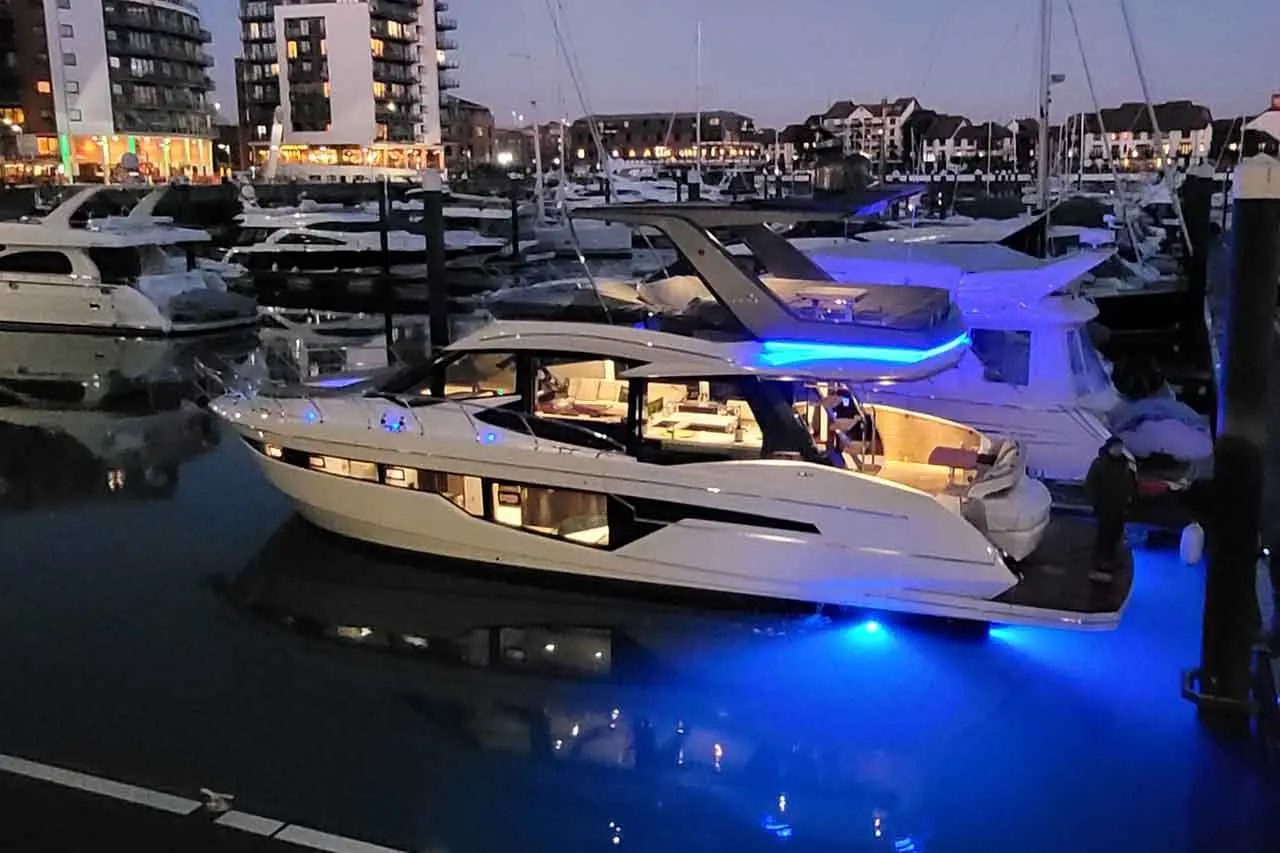 Luxury yacht illuminated with blue lights docks at a marina, surrounded by other boats and modern buildings at dusk.