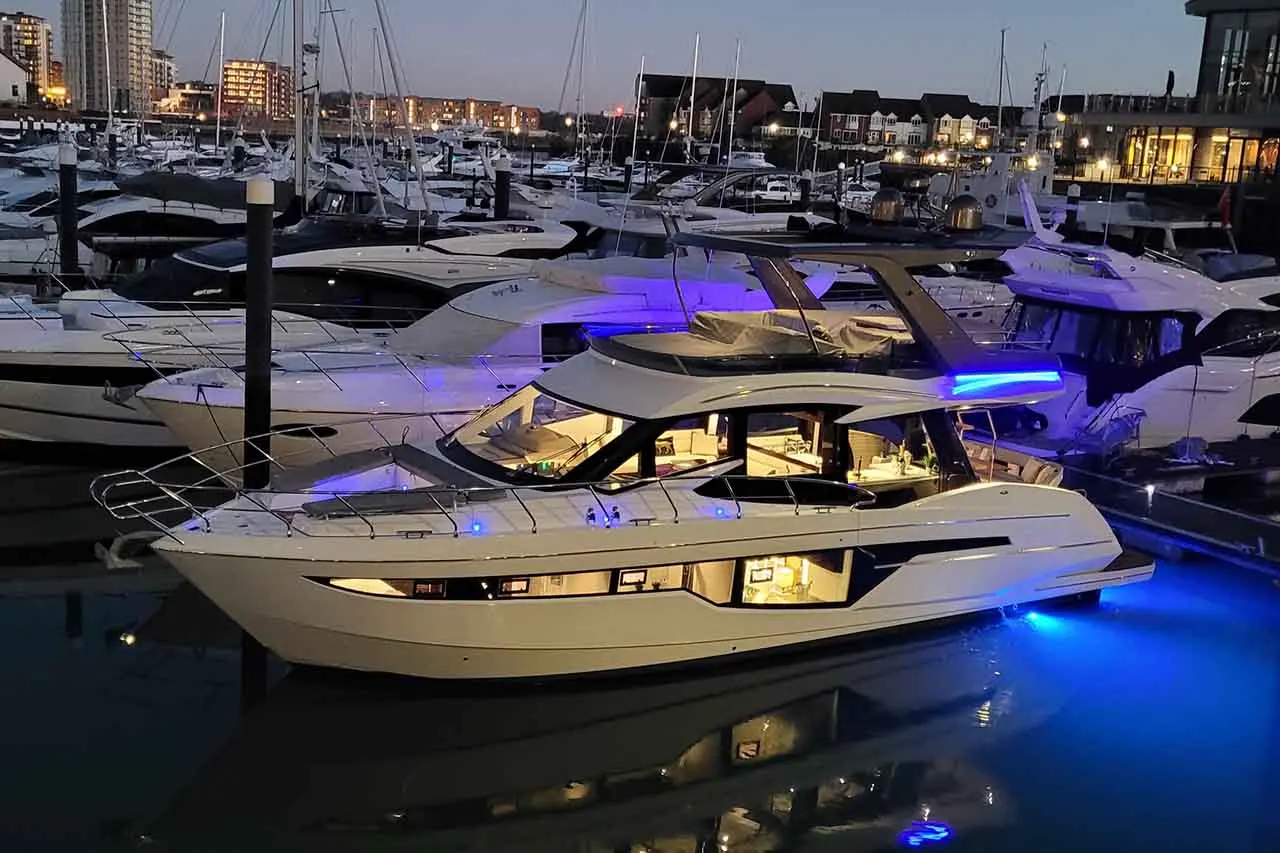 Luxury yacht illuminated with blue lights docked in a marina, surrounded by other boats and city buildings at dusk.